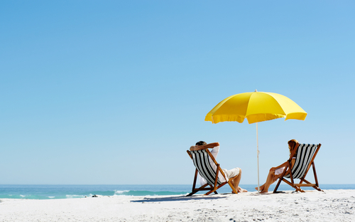 Sandy beach with two people sat on deckchairs beneath a parasol
